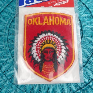 May include: An iron-on patch featuring the word "Oklahoma" above an image of a Native American person wearing a headdress. The patch is red with yellow and white accents, and is packaged in a clear plastic sleeve. The patch is labeled "Patches" and "Made in U.S.A. by Voyager".