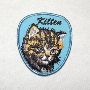 Könnte beinhalten: Gestickter Aufnäher mit einem Kätzchen mit braunem, schwarzem und orangefarbenem Fell, vor hellblauem Hintergrund. Das Wort "Kitten" ist in Schreibschrift über dem Kopf der Katze gestickt. Der Aufnäher hat einen blauen Rand.