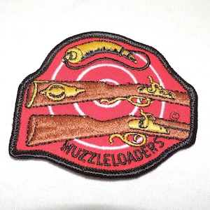 Puede incluir: Un parche bordado rojo y negro con dos armas de avancarga y un blanco. El texto "MUZZLELOADERS" está escrito en negro en el parche.
