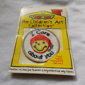 May include: A white, round embroidered patch with a yellow smiley face with red hair and the text "I Care about you". The patch is on a yellow card with the text "Voyager presents the Children's Art Collection".