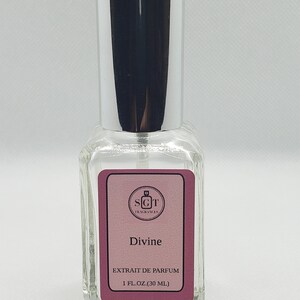 May include: A clear glass bottle with a silver cap and a pink label. The label says "Divine" and "Extrait de Parfum" with the brand name "SGT Fragrances" in a small square at the top. The bottle contains 1 fl oz (30 ml) of perfume.