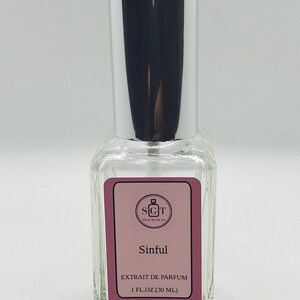 May include: A clear glass bottle with a silver spray top and a pink label. The label says "SGT Fragrances" and "Sinful" in white text. It also says "Extrait de Parfum" and "1 fl. oz. (30 ml)" in white text.