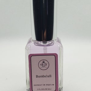 May include: A clear glass bottle with a silver cap and a pink label. The label says "SGT Fragrances" and "Bombshell" in black text. The label also says "Extrait de Parfum" and "1 fl. oz. (30 ml)" in smaller black text.