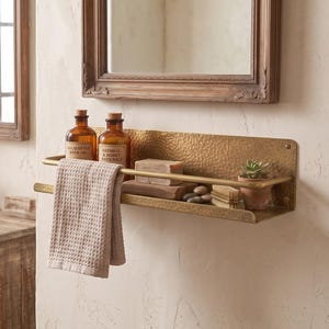 May include: A wall-mounted brass bathroom shelf with a hammered texture, featuring two horizontal bars. The shelf holds two amber glass bottles, a bar of soap, a small potted succulent, and a beige waffle-weave towel draped over the front rail.