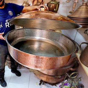 Puede incluir: Olla de cocina grande de cobre martillado con tapa a juego. La olla es de forma ovalada con un borde ancho y asas. La tapa tiene una parte superior redondeada con un pequeño asa. El cobre tiene un color marrón rojizo cálido y una superficie texturizada.