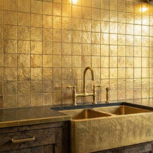 May include: A kitchen area featuring a double-basin hammered brass farmhouse sink with a matching brass bridge faucet. The backsplash consists of square hammered brass tiles, and the dark wood cabinetry is topped with a dark stone countertop.
