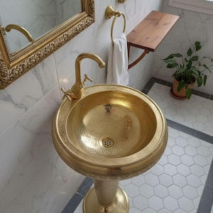 May include: A gold-colored pedestal sink with a hammered texture and intricate engraved patterns on the basin and base. It features a matching gold-colored curved faucet with two cross-style handles and a circular drain at the center of the basin.