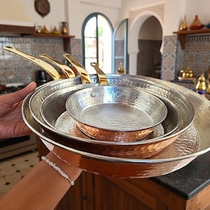 May include: Set of four hammered copper and brass pans with gold-colored handles. The pans are nested together, with the smallest pan at the top and the largest at the bottom. Kitchenware.
