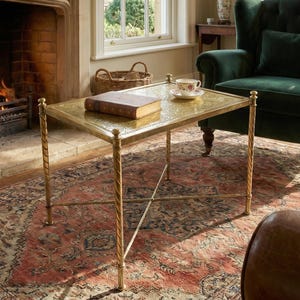 Puede incluir: Salón con una mesa de centro dorada, un libro y una taza de té. La mesa está sobre una alfombra estampada, con un sillón verde y una chimenea con fuego. La habitación tiene un ambiente tradicional y acogedor.