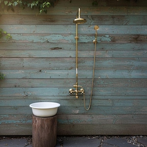 Può includere: Doccia esterna con finitura dorata, montata su una parete in legno blu-verde invecchiato. Un lavabo bianco poggia su un ceppo di legno. Il fogliame verde incornicia la scena, creando un'estetica naturale e rustica.