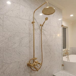 May include: A gold shower system featuring a round overhead shower head and a handheld shower wand mounted on a white marble tiled wall. The fixture includes a central control valve with two handles and a curved pipe design in a metallic finish.