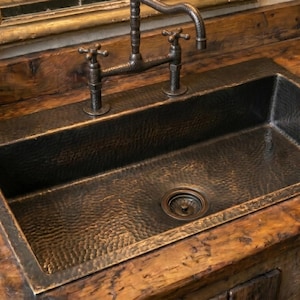 Könnte beinhalten: Ein gehämmertes Kupferbecken mit dunkler Oberfläche ist in eine rustikale Holz-Arbeitsplatte eingelassen. Das Waschbecken hat einen zentralen Abfluss und wird mit einem Wasserhahn im Vintage-Stil kombiniert. Ein geflochtener Korb mit gerollten Handtüchern steht daneben.