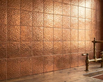 Puede incluir: Una pared cubierta con azulejos de cobre cuadrados y martillados con acabado metálico. Un grifo de metal color latón con dos manijas y un caño curvo está montado frente a los azulejos, sobre una encimera de madera y un fregadero de cobre parcial.