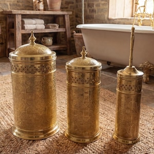 Puede incluir: Tres accesorios de baño dorados: un cubo de basura alto con tapa, un recipiente más pequeño con tapa y un portacepillos de inodoro. Todas las piezas tienen un diseño en relieve adornado. Los artículos están sobre una alfombra tejida en un baño.