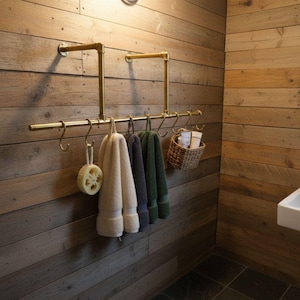Puede incluir: Interior de baño con toallero de latón con toallas crema, grises y verdes. También hay una esponja vegetal natural y una pequeña cesta de mimbre. La pared es de tablones de madera, con lavabo y espejo.