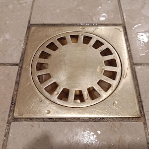 Könnte beinhalten: Ein quadratischer, metallischer Duschablauf mit einem kreisförmigen, geschlitzten Deckel. Der Abfluss ist in helle Fliesen verlegt, wobei Wassertropfen auf der Oberfläche sichtbar sind. Das Design des Abflusses ist einfach und funktional.