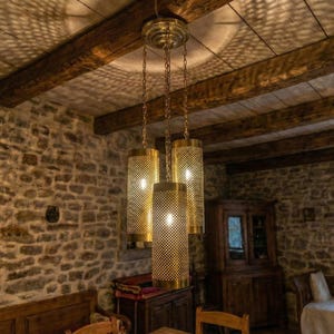 May include: A brass chandelier with three cylindrical, perforated shades hangs from a wooden beam ceiling. The light casts a patterned shadow on the ceiling. The room has stone walls and wooden furniture, creating a rustic ambiance.