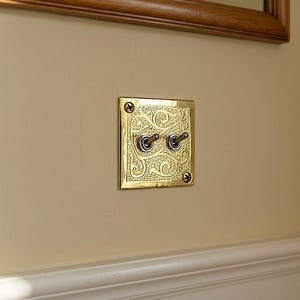 May include: A brass light switch plate with two toggle switches. The plate features an ornate, swirling design and is mounted on a beige wall. The switches are in the on position.