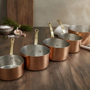May include: A set of five copper saucepans with brass handles, arranged on a wooden surface. The pans have a hammered texture and a shiny interior. Steam rises from one of the pans, suggesting it is in use.