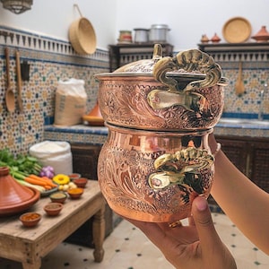 Pode incluir: Panela de cozinha de cobre com desenhos florais e de folhas ornamentados. A panela tem tampa e alças decorativas. A imagem mostra um ambiente de cozinha com uma mesa de madeira, um tajine e várias especiarias.