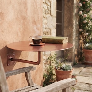 Puede incluir: Una mesa redonda de pared en color cobre con superficie texturizada, fijada a una pared de estuco mediante un brazo metálico. Una taza de cerámica con plato y un libro de tapa dura cerrado están colocados sobre la mesa, con vapor saliendo.