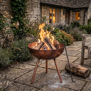 May include: A round, hammered copper fire pit with three slender legs, containing a burning wood fire with bright orange flames. A long metal fire poker rests beside the bowl, and a single log lies on the stone ground near a wooden chair.