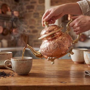 May include: A copper teapot with ornate detailing pours tea into a speckled ceramic mug. The scene includes a wooden tea box, additional mugs, and a vase with flowers, all set on a wooden table. Steam rises from the tea, suggesting warmth.