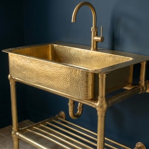 Puede incluir: Un lavabo de latón martillado con un grifo a juego. El lavabo rectangular se asienta sobre un marco de tubería de latón con un estante inferior. El accesorio está contra una pared azul oscuro.