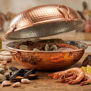 Puede incluir: Una campana de cobre con mariscos en su interior, incluyendo langosta, almejas y camarones. La campana está abierta, revelando el contenido. Otros ingredientes incluyen pescado, limones, perejil, ajo y una botella de aceite de oliva. También hay una copa de vino blanco.