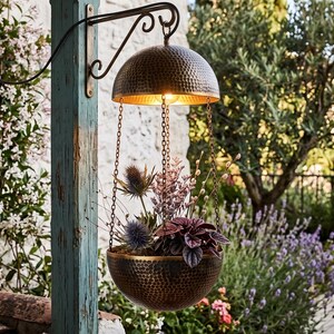 May include: A hanging planter featuring a hammered bronze-tone metal bowl and a matching dome top connected by three thin chains. The planter contains various plants including purple thistle and dark foliage, suspended from a black metal wall bracket.