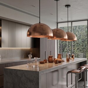 May include: Three large, dome-shaped copper pendant lights with a hammered texture hang above a kitchen island. The island features a gray stone countertop with various glass decanters, wooden bowls, and two stools with brown leather seats.