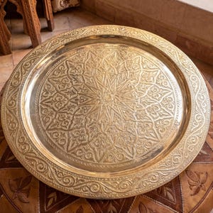 Könnte beinhalten: Ein runder, goldfarbener Metalltablett mit aufwendigen geätzten Mustern, das auf einem braunen Lederhocker liegt. Das Design des Tabletts zeigt einen zentralen Stern und einen floralen Rand, was einen dekorativen Stil zeigt.