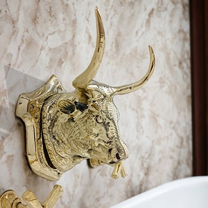 May include: A gold bull head sculpture mounted on a wall, with a faucet coming out of its mouth. The bull's head is detailed with textured fur and horns.