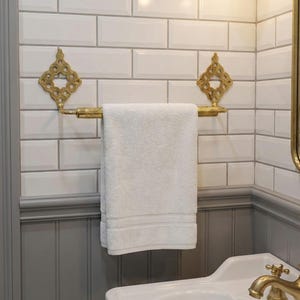 May include: A gold-toned towel rack with a white towel hanging on it. The rack has decorative diamond-shaped accents. The background features white subway tiles and gray wainscoting, creating a classic bathroom aesthetic.
