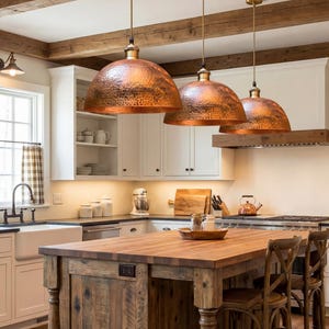 Puede incluir: Una cocina con gabinetes blancos, una isla de madera y tres lámparas colgantes de cobre. La isla tiene una encimera de bloque de carnicero y sillas de madera. La cocina también cuenta con una ventana con cortina a cuadros y estanterías abiertas.