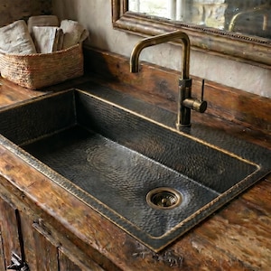 May include: A rustic bathroom sink with a hammered copper basin and a bronze faucet. The sink is set into a weathered wooden countertop and cabinet. A woven basket with folded towels sits nearby.