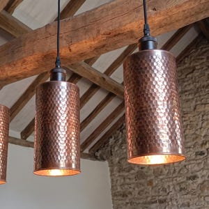 Peut inclure: Trois suspensions cylindriques avec une finition en cuivre martelé. Chaque luminaire est suspendu par un cordon noir et présente une surface texturée et alvéolée qui reflète la lumière. Le bas ouvert de chaque cylindre révèle une ampoule chaude.