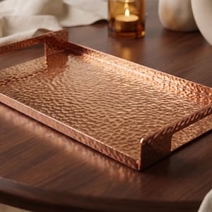May include: A rectangular copper-colored serving tray with a hammered, textured surface and integrated handles on both ends. The tray features a metallic finish and raised edges, providing a flat base for holding items on a flat surface.