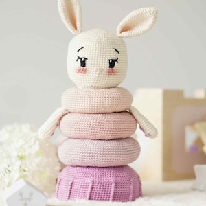 Puede incluir: Un juguete apilable de conejito de ganchillo hecho a mano. El conejito tiene una cabeza color crema con mejillas rosadas y ojos bordados en negro. Los anillos apilables son tonos de rosa y morado. El juguete está diseñado para bebés y niños pequeños.
