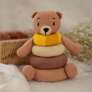 Può includere: Un orsacchiotto di peluche marrone all'uncinetto con un giocattolo ad anelli impilabili giallo, beige e marrone. L'orso ha occhi neri e un naso nero.