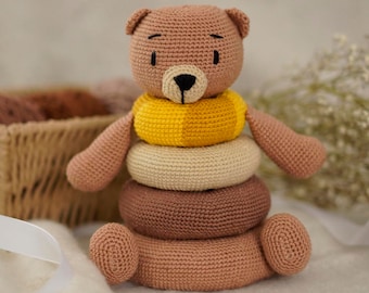 May include: A crocheted brown bear toy with a yellow, beige, and brown stacking ring toy. The bear has black eyes and a black nose.