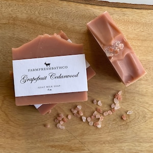 May include: Two bars of pink goat milk soap with a white label that reads "Farm Fresh Bath Co. Grapefruit Cedarwood - Goat Milk Soap - 4 oz". The soap is on a wooden surface with pink Himalayan salt scattered around it.