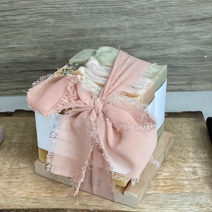 May include: A stack of handmade soaps, wrapped with a light pink ribbon and bow. The soaps are various shades of green and yellow, with visible natural ingredients. The label reads "Farmer Lemon". The soaps rest on a wooden soap dish.