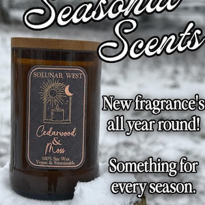 May include: A brown glass candle with a wooden lid, labeled "Cedarwood & Moss" with a black and gold label. The text "Seasonal Scents" is at the top. Additional text reads "New fragrance's all year round!" and "Something for every season."