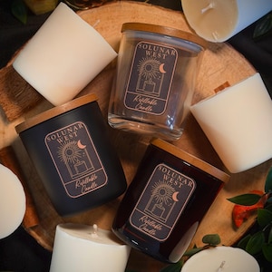 May include: Three glass and matte black candle jars with wooden lids, each featuring a label with the text Solunar West Refillable Candle and a sun and moon graphic. They are surrounded by several white wax candle refills without containers.