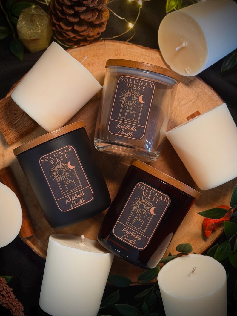 May include: Three glass and matte black candle jars with wooden lids, each featuring a label with the text Solunar West Refillable Candle and a sun and moon graphic. They are surrounded by several white wax candle refills without containers.