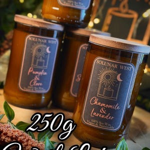 May include: Several glass jars of scented candles with wooden lids are stacked. Each jar has a label with the brand name "SOLUNAR WEST" and a different scent name, such as "Pumpkin & Clove" and "Chamomile & Lavender". The text "250g With Lid" is also visible.