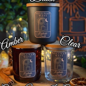 May include: Three refillable candle jars with wooden lids, labeled Solunar West. The jars come in black, amber, and clear glass. Each jar features a rectangular label with a sun and moon graphic. The text reads Choose Your Jar Colour.