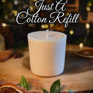 May include: A cylindrical white candle refill with a centered cotton wick. The text "Just A Cotton Refill" is displayed in a white cursive font with a black outline above the candle. The candle has a smooth, solid, opaque wax surface.