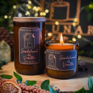 May include: Two glass jar candles with wooden lids, labeled "Sandalwood & Vanilla" by Solunar West. The candles are on a wooden surface with dried orange slices and greenery. One candle is lit, showing a flame.